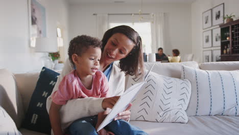 mamá en el sofá en la sala de estar leyendo al niño sentado en su rodilla, papá e hija en el fondo