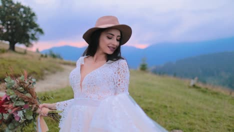 Smiling bride in beautiful white dress. Young woman in hat waving her wedding dress on the nature background in the evening. Girl with bouquet of wild flowers.