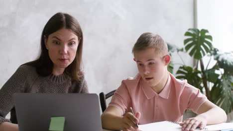 Close-up-view-of-a-boy-with-down-syndrome-watching-something-on-laptop-sitting-at-table-with-his-mother-in-the-living-room-at-home