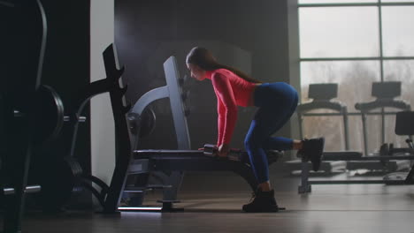 La-Mujer-Con-Un-Top-Rosa-Corto-Está-Trabajando-Boca-Arriba-En-El-Gimnasio.-Se-Apoya-En-Un-Banco-Con-La-Parte-Derecha-Del-Cuerpo-Y-Entrena-La-Mano-Izquierda-Y-La-Parte-Izquierda-De-La-Espalda.