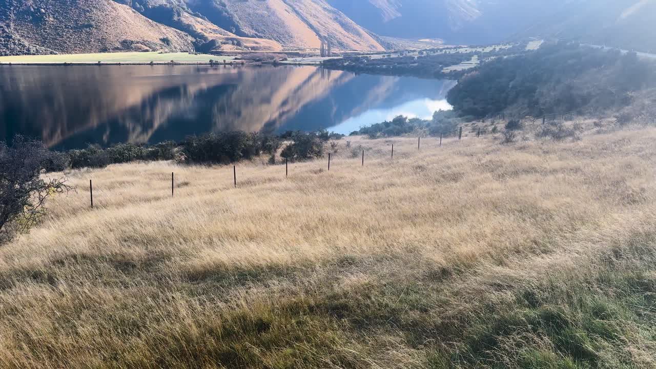 Camera rises from grassy field to reveal Lake Moke, mountains, and blue sky in daylight