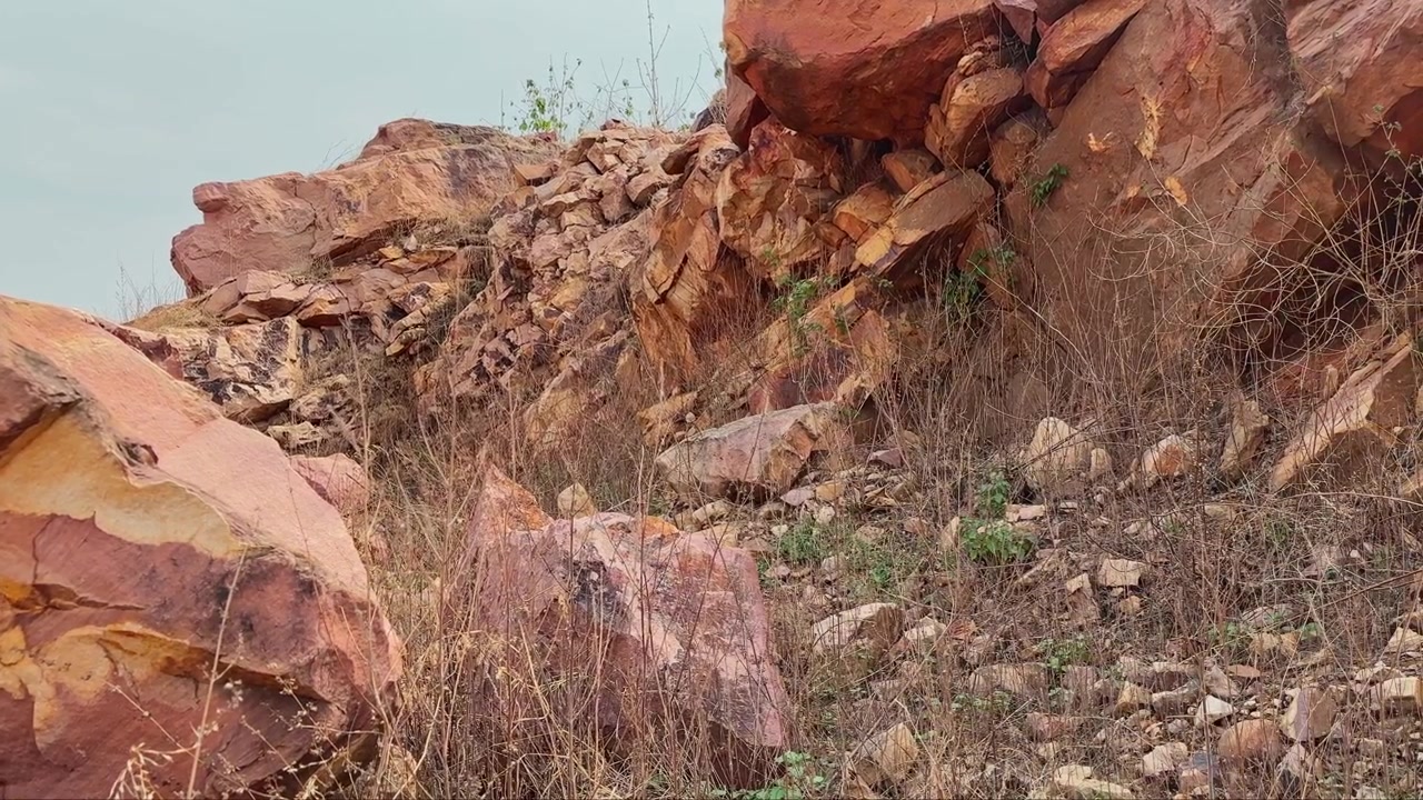 Lenta carrellata in avanti verso un aspro affioramento roccioso caratterizzato da massi fratturati, superfici di pietra erose e vegetazione di erba secca. Texture naturali e formazioni ricche di minerali dominano il paesaggio