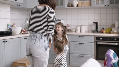 madre divertida, joven y alegre está jugando con sus hijas. imitando montar a caballo usando palas en la cocina mientras cocina