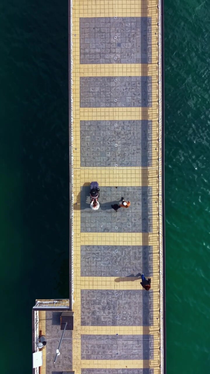 Persone camminano su un ampio molo con un pavimento giallo e grigio lungo l'acqua in una giornata limpida all'aperto.