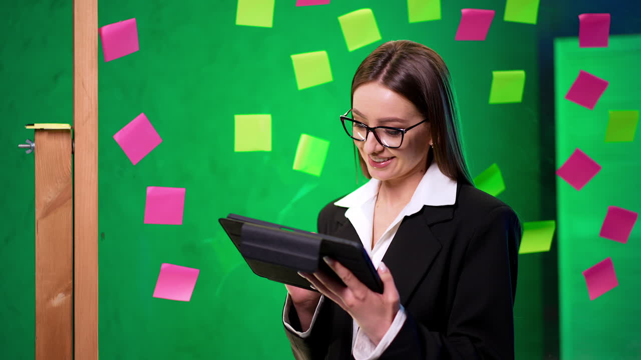 Beautiful smiling brunette lady in glasses and jacket holds a tablet. Glass wall with sticky notes at backdrop.
