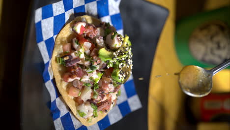 vertical slow motion of a seafood tostada with avocado and sesame on top placed on a blue checkered paper and someone pouring green salsa sauce on top