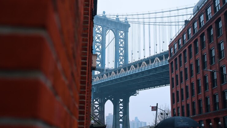 Architectural detail of suspension bridge framed by red brick wall. Close up view of Manhattan Bridge tower through a narrow opening between brick buildings in Dumbo area