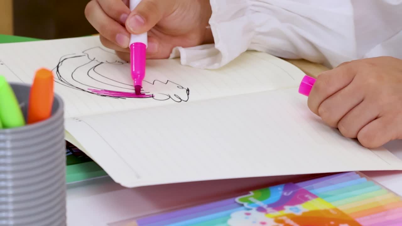 幼い生徒がピンクのフェルトペンで、ノートに虹の色を塗っている。