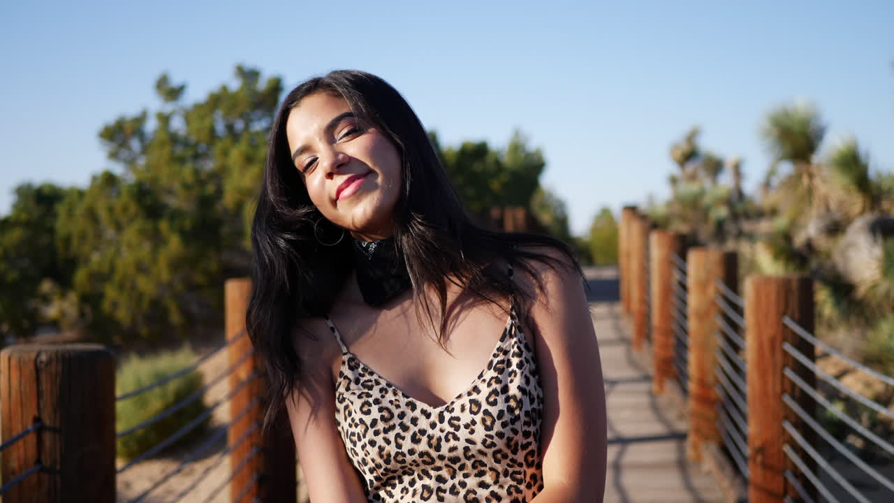 Cute young hispanic woman traveler walking in slow motion along a wooden path outdoors in sunlight smiling and looking happy