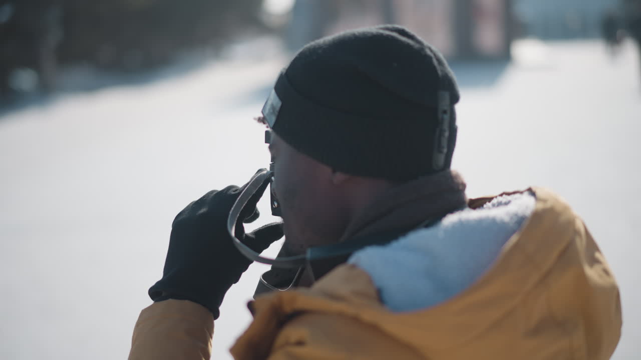 wildlife photographer in yellow parka and black beanie peering through vintage camera viewfinder with gloved hands fine tuning focus dial against bright urban winter sky over snowy plaza