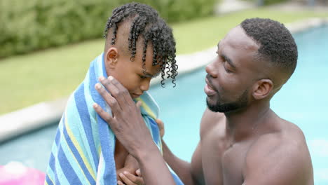 Un-Padre-Afroamericano-Envuelve-Una-Toalla-Azul-Alrededor-De-Su-Hijo-Junto-A-La-Piscina-En-Casa