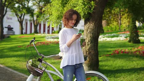 Portrait-of-a-young-woman-typing-a-message-using-her-smartphone-standing-in-the-city-park-near-her-city-bicycle-with-flowers-in