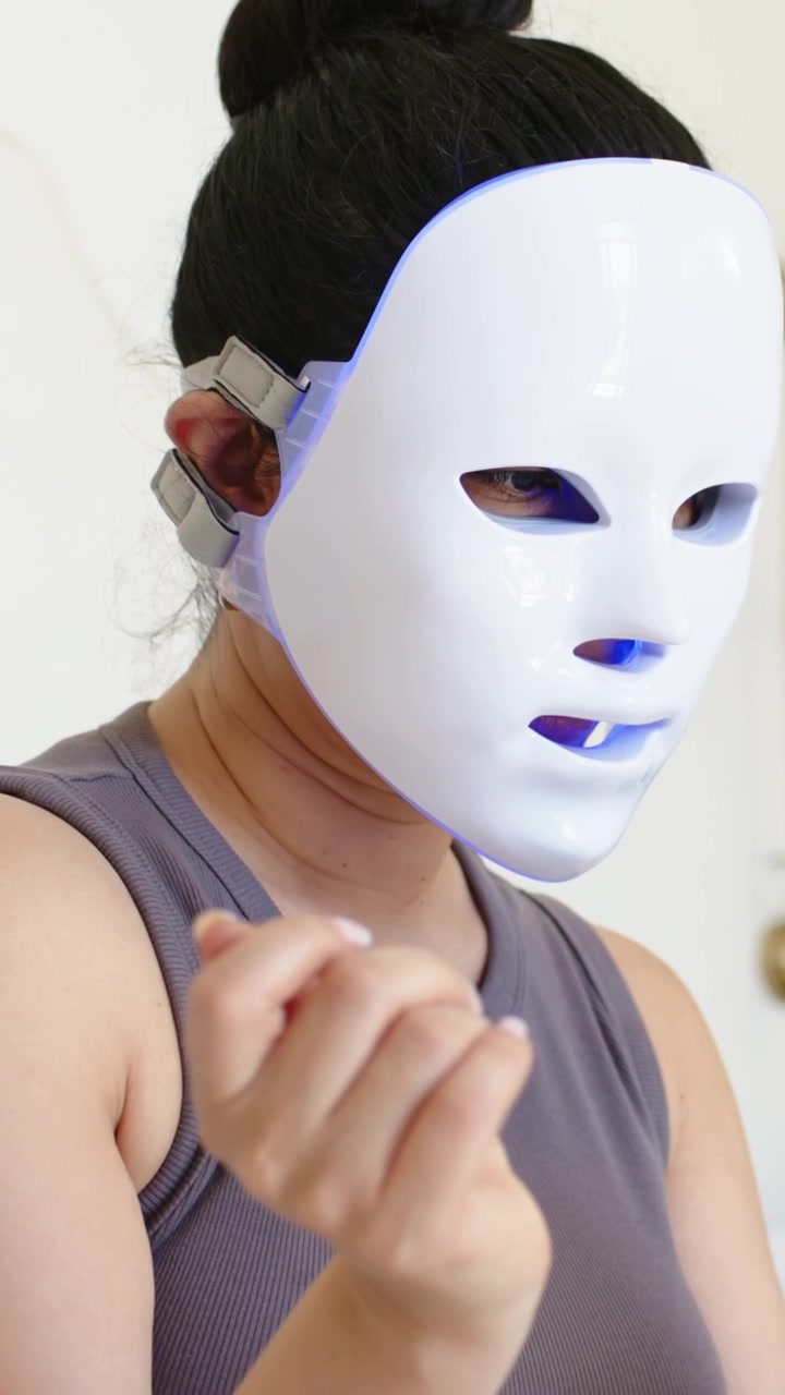 Vertikal video: Siddende kvinde iført grå tanktop og LED-maske ved sminkebordet, justerer og tapper derefter på telefonen.