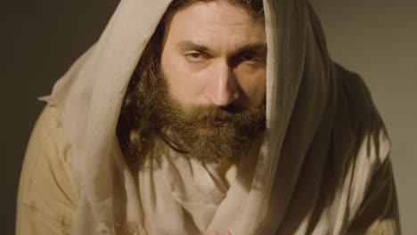 man wearing robes with long hair and beard representing figure of jesus christ looking up into camera and praying