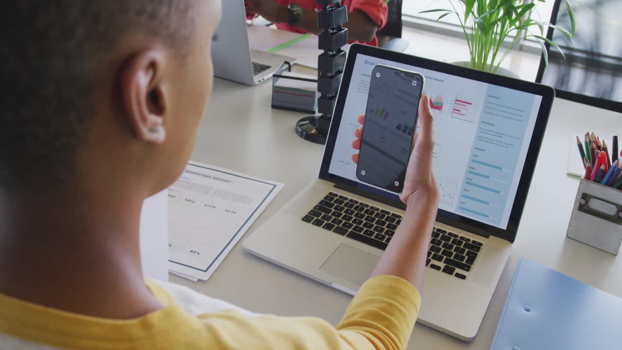 African american businesswoman at desk, reading report, using laptop and smartphone with copy space