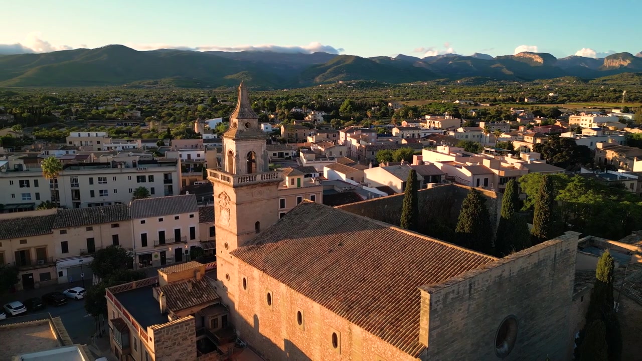 Vista aerea della chiesa di Santa Maria del Camí circondata da un incantevole villaggio mediterraneo a Maiorca, Spagna, con montagne distanti e vegetazione sotto un cielo sereno.