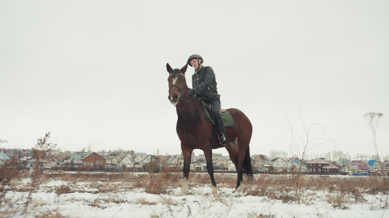 Cavaliere in sella in un campo aperto innevato che pratica postura ed equilibrio durante una sessione di educazione equestre. L'equestre con il casco siede eretto su un cavallo marrone, mentre la rada vegetazione invernale e le case distanti incorniciano la scena.