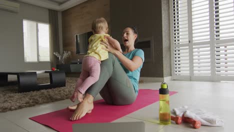 caucasian mother playing with her baby while practicing yoga on yoga mat at home