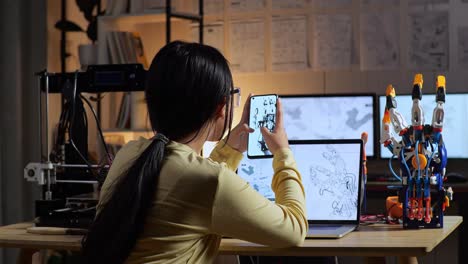 back view of teen asian girl with 3d printing looking at the pictures on smartphone while designing a cyborg hand on a laptop at home