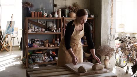 Portrait-Of-Female-Potter-Wearing-Beige-Apron-Putting-Clay-Piece-On-Worktop-And-Then-Starting-Kneading-It-With-Her-Hands