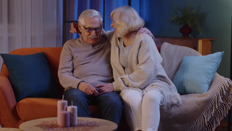 Happy-old-senior-elderly-family-couple-hugging,-laughing,-smiling-talking-at-modern-home-sofa