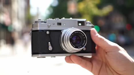 a person holds a classic camera while standing on a bustling city street. sunlight shines down, creating a vibrant atmosphere filled with energy and urban life.