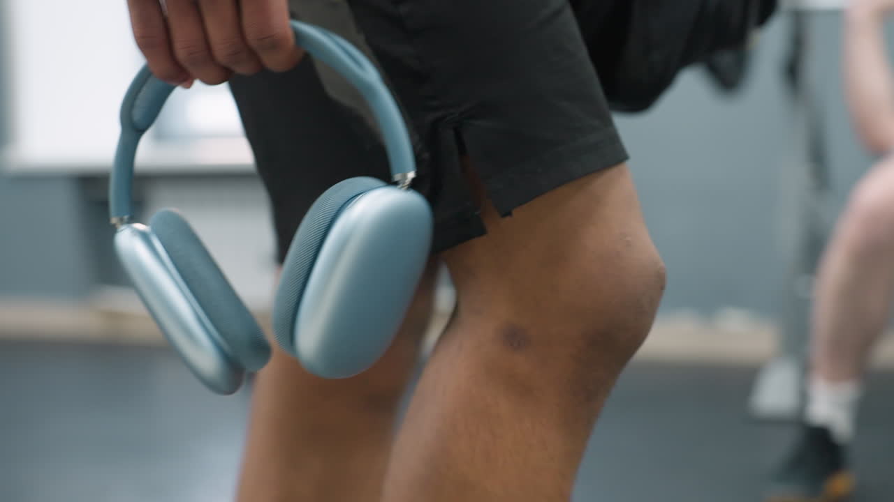close up lower body view of person in black shorts and white sneakers walking past treadmill in modern gym while holding light blue headphones by side