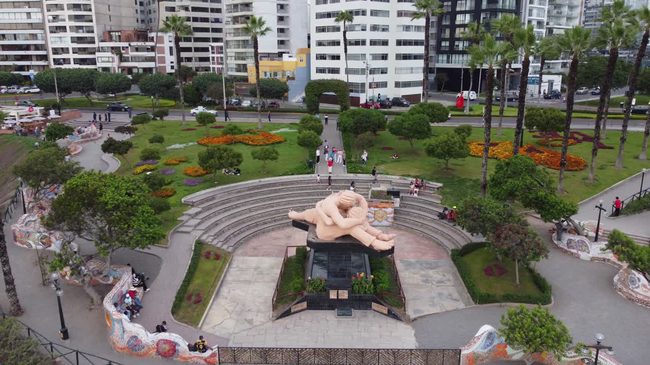 Drone flies over a park with sculpture of couple kissing. Many people sit and walk around the park surrounded green grass and trees. Located in "Parque del amor" in Miraflores district of Lima, Peru.