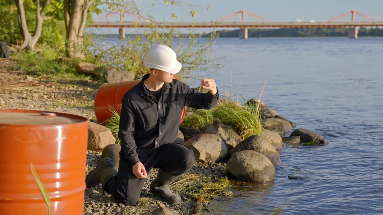 Scientist kneels near lake edge collecting and swirling water into sample bottle during test near oil drum barrels