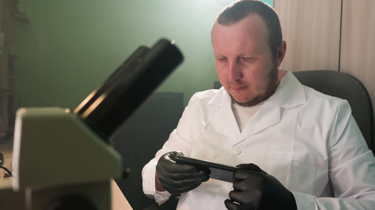 백색 실험실 코트와 검은 장갑을 입은 기술자가 실험실의 책상에 앉아 스마트폰을 조심스럽게 조사하는 클로즈업 뷰