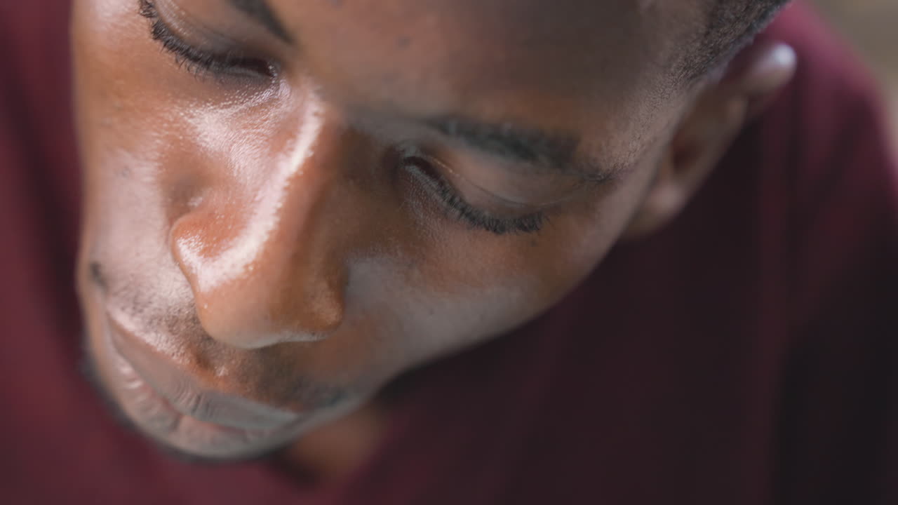 Close-up de homem negro estudando, camisa bordô, gotas de suor na testa, olhar cabisbaixo, profundidade de campo rasa, iluminação suave e quente, expressão introspectiva e concentração silenciosa em estúdio espontâneo.