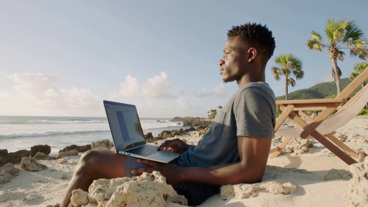 Homem digitando vestindo camiseta cinza, shorts na praia pausando enquanto as ondas chamam a atenção, laptop no colo