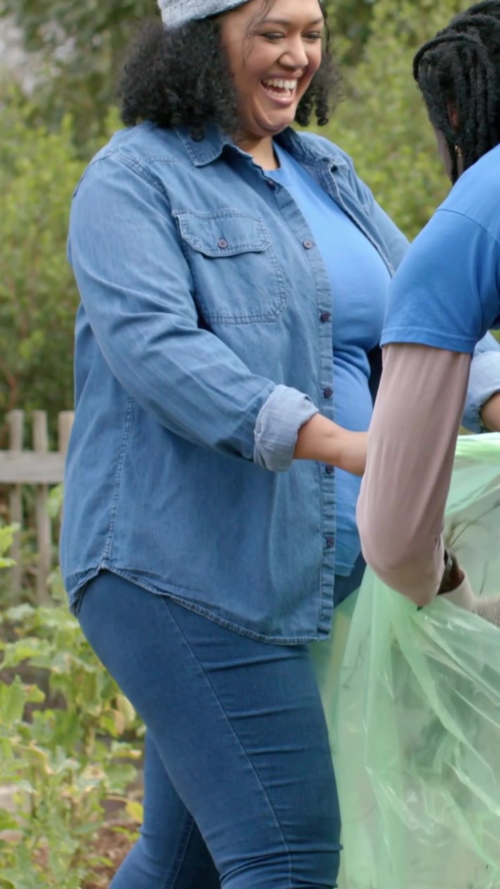 Vertical video: Arriving with green bag urging blue tee adding weeds into bag at garden for cleanup