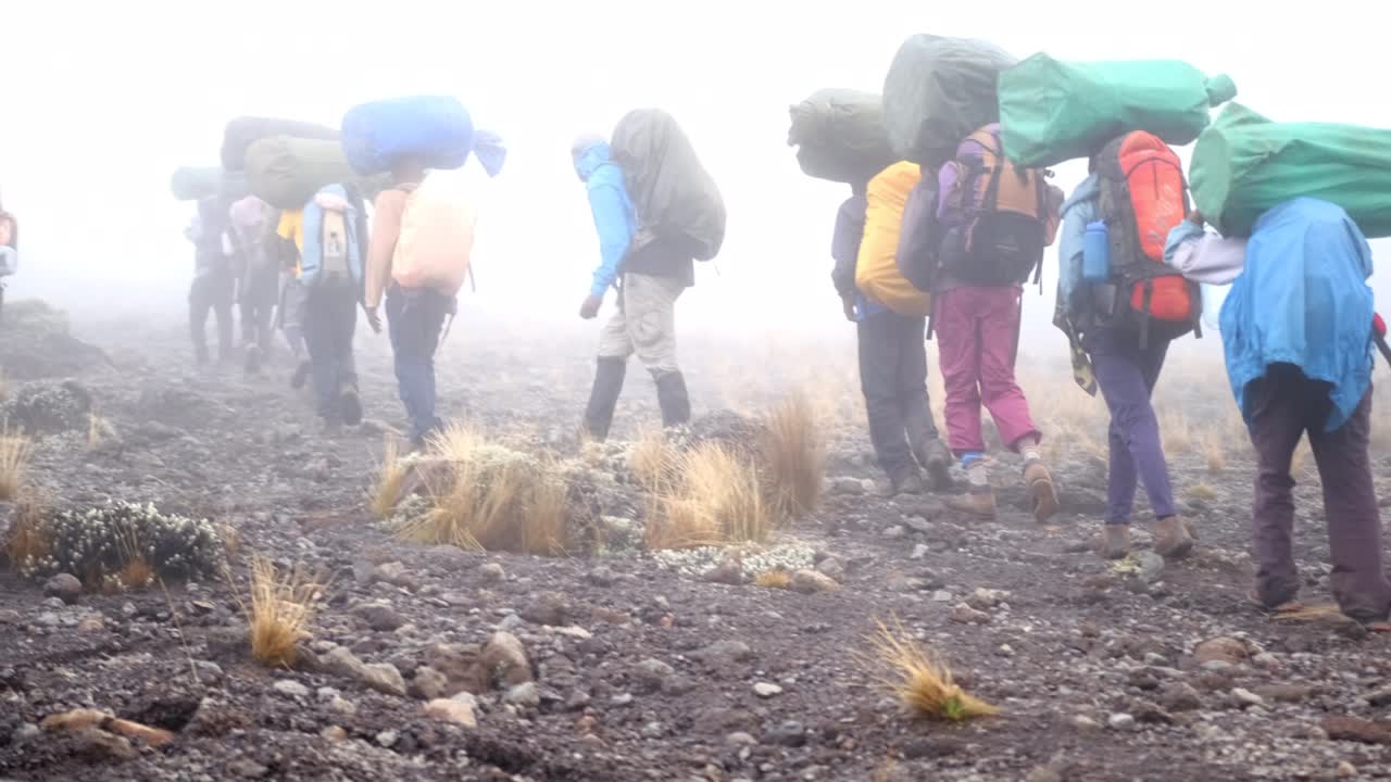 A group of porters with trunks on their heads and tourists with backpacks