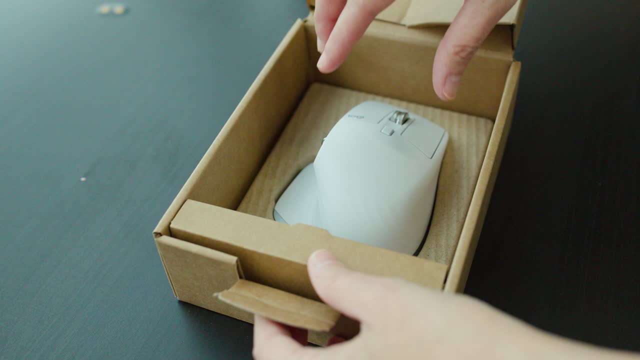 Hands open cardboard box, remove white wireless mouse, natural daylight, close-up, static camera