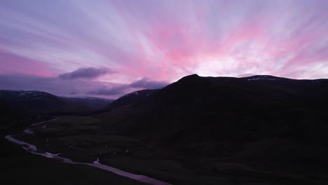 aerial drone footage of glenshee in scotland during an intense pink and purple sunset looking up the glen towards the silhouetted mountains as the sunset reflects in a river and off of the clouds