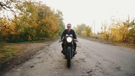 Front-view-of-a-stylish-cool-young-man-in-sunglasses-and-leather-jacket-riding-motorcycle-on-a-asphalt-road-on-a-sunny-day-in