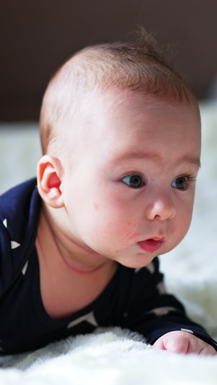 Little newborn baby looks puzzled gazing aside. Beautiful boy lies on belly keeping head up. Blurred backdrop. Vertical video