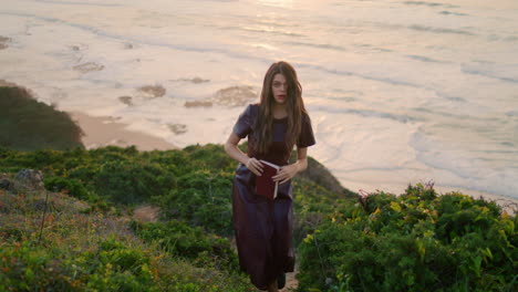 woman holding book walking green hill in front evening ocean. girl rest seashore