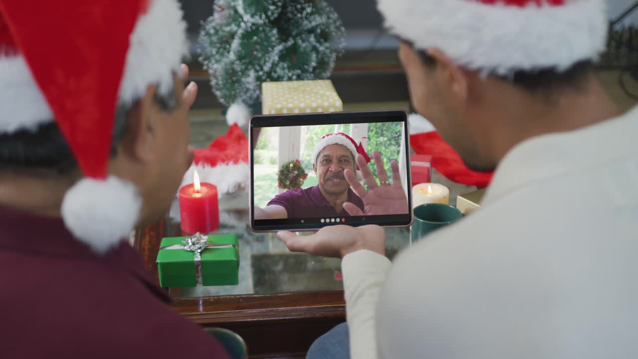 padre y hijo biraciales agitando y usando la tableta para la videollamada de navidad con el hombre en la pantalla
