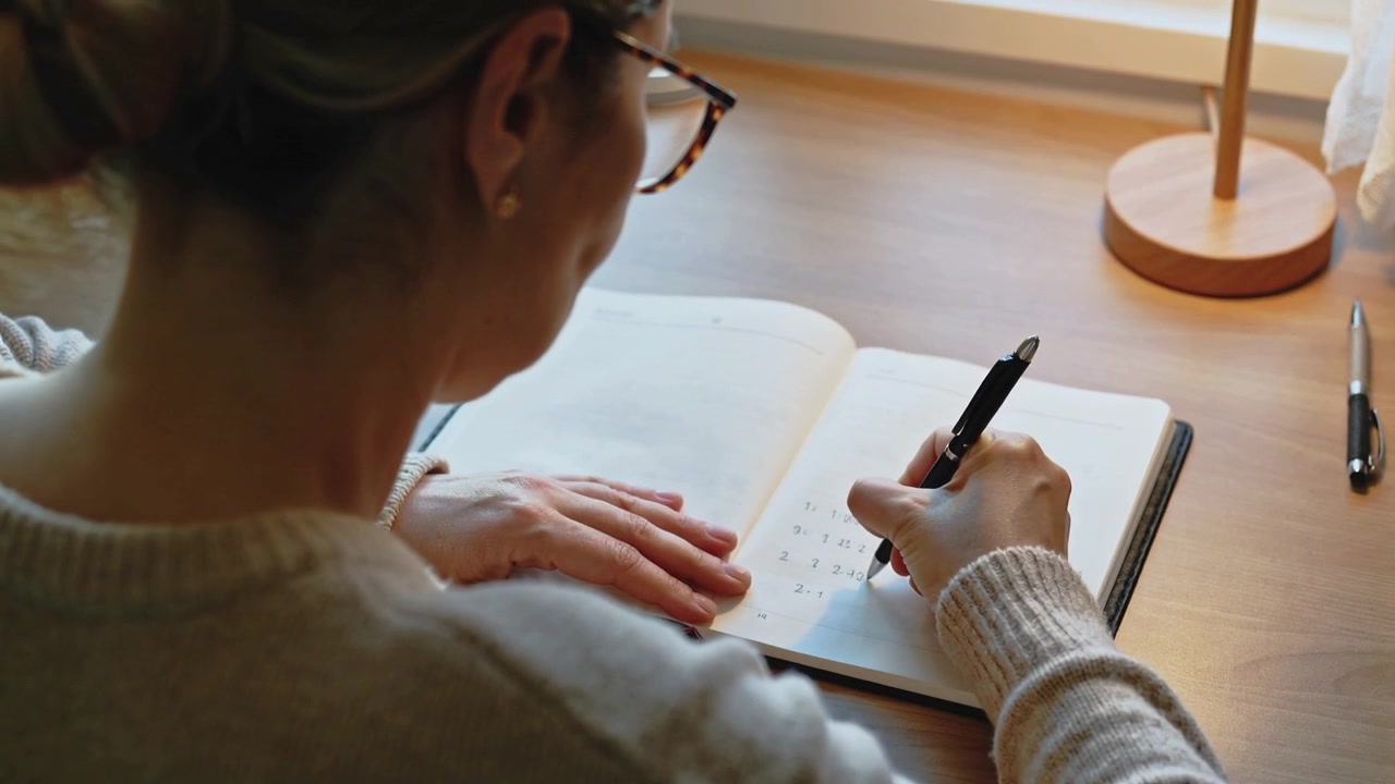 Writing woman with bun, glasses starting notebook list for notes at desk by window, holding pen