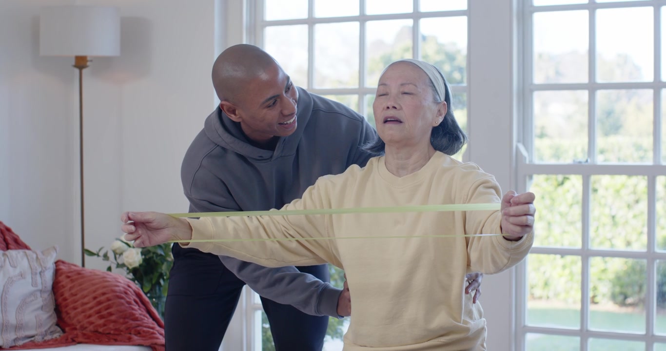 Carer assisting Senior Asian woman holding band at home approaching adjusting supporting posture