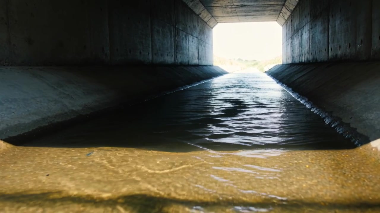 Acqua che scorre entra da un'apertura illuminata dal sole, muovendosi verso la telecamera all'interno di un canale scatolare con un letto sabbioso