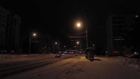 Einsame-Bushaltestelle-In-Der-Nacht-Auf-Einer-Verschneiten-Straße,-Beleuchtet-Von-Warmen-Straßenlaternen-Und-Vorbeifahrenden-Autos-Unter-Dem-Schneefall