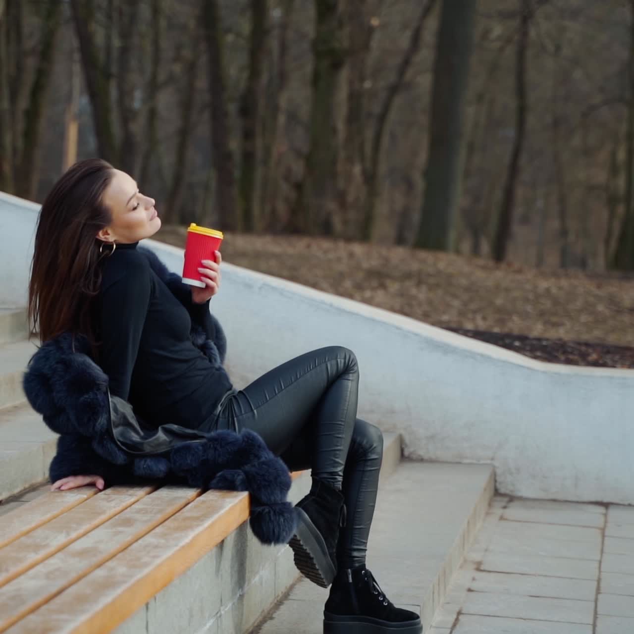 Sexy young woman with a cup of coffee outdoors. Beautiful lady sitting alone on the bench in the park in cold season and drink tea. Slow motion.