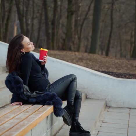 sexy young woman with a cup of coffee outdoors. beautiful lady sitting alone on the bench in the park in cold season and drink tea. slow motion.