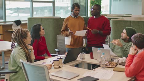 Treffen-Von-Kollegen,-Die-An-Einem-Tisch-Im-Büro-Sitzen,-Zwei-Kollegen-Stehen-Und-Sprechen-über-Ein-Projekt