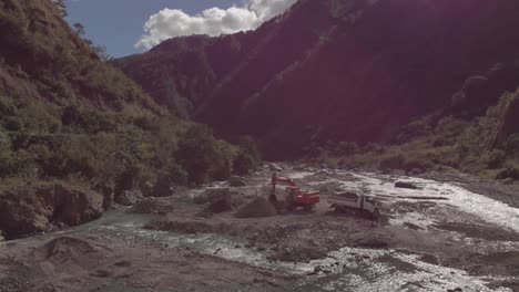 excavator digging rocks out of river bed in mountainous region in Benguet Kabayan Philippines wide angle aerial approaching raw cement material mining flowing water reflecting light bright sunny day