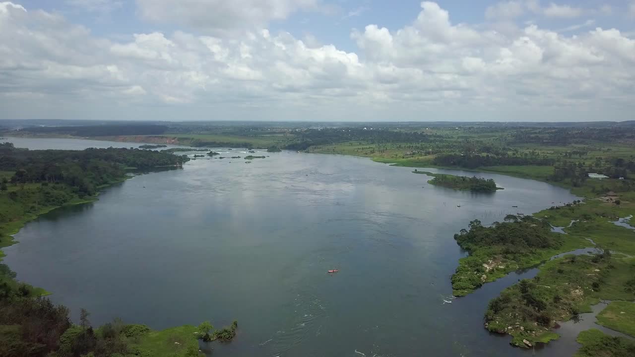 Aerial drone footage pulling away over the majestic River Nile in Uganda, showing calm blue waters bordered by lush green vegetation, distant farmland, and a partly cloudy tropical sky