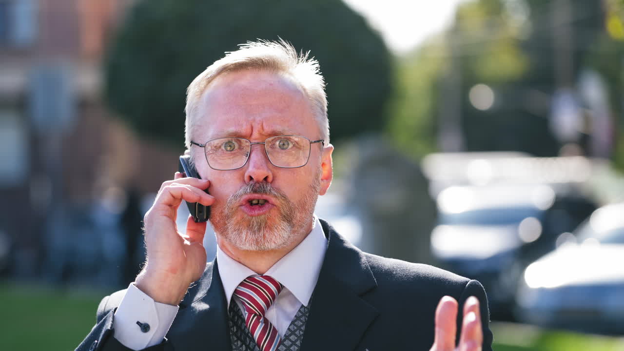 Close-up face of elderly man in eyeglasses talking on cellphone outdoors. Portrait of a handsome senior bearded businessman against city background.